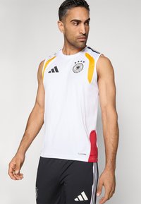 Maiou sportiv alb fără mâneci, cu accente negre și logo-ul federației germane de fotbal. Prezintă panouri laterale portocalii și roșii, confecționate din țesătură texturată.