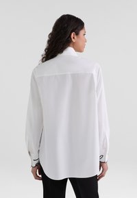 White shirt with long sleeves, curved hem, and black accent stitching at cuffs. Smooth texture and standard collar design.