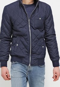 Mężczyzna w granatowej pikowanej kurtce na zamek błyskawiczny z kieszeniami na zatrzaski i jasnoniebieskich dżinsach, stojący na jednolitym tle.