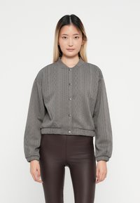 Veste bomber en tricot gris avec col rond, fermeture à boutons et poignets élastiques, associée à un pantalon ajusté marron. Texture lisse.
