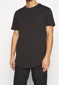 T-shirt in cotone nero con scollo rotondo e maniche corte, caratterizzato da un orlo curvo. Il tessuto appare morbido con una superficie liscia.