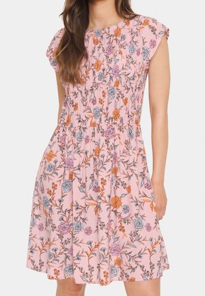 Kobieta ubrana w różową sukienkę z krótkim rękawem w pomarańczowo-niebiesko-fioletowy wzór kwiatowy, z marszczoną talią i rozszerzanym dołem.