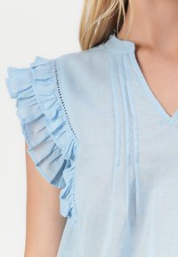 Blusa de algodón azul claro con mangas acampanadas, textura de puntos, escote en V y pliegues verticales a lo largo del frente, que añaden un detalle sutil.