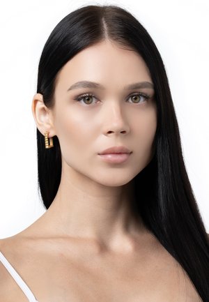 Goldene Creolen mit strukturiertem Design, getragen von einem Model mit langen, glatten schwarzen Haaren und einem neutralen Makeup-Look vor einem weißen Hintergrund.