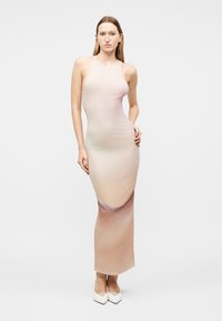 Γυναίκα στέκεται φορώντας ένα αμάνικο, εφαρμοστό, μακρύ φόρεμα σε μπεζ χρώμα με λευκά παπούτσια με μυτερή μύτη, μπροστά σε απλό λευκό φόντο.