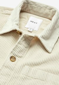 CremefarbigesCord-Hemd mit kragenförmigem Ausschnitt, zwei Knöpfen und einer Brusttasche. Verfügt über eine vertikale Rippenstruktur und ein Markenetikett innen.