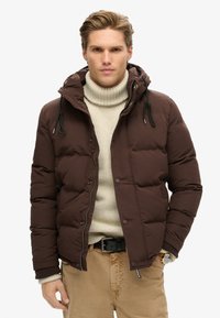 Chaqueta acolchada marrón con capucha forrada de polar, cierre con cremallera y a presión, llevada sobre un cuello de tortuga crema. Presenta puños elásticos y cordones ajustables.