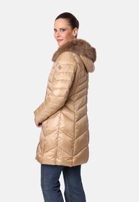Mujer vestida con un abrigo acolchado beige con capucha de piel, de pie en un perfil tres cuartos frente a un fondo liso.