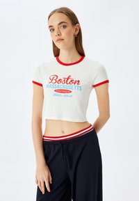 Hvid cropped t-shirt med rød og blå tekst, der viser "Boston Massachusetts" design. Korte ærmer med rød kant. Matchet med navy bukser.