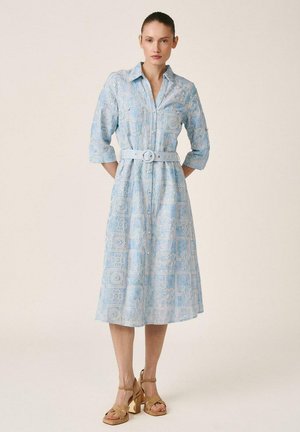 Mujer con un vestido midi azul claro con patrón floral de encaje, mangas remangadas, cintura ceñida con cinturón y sandalias de tacón alto color marrón claro, posando contra un fondo liso.