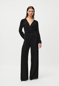 Czarny kombinezon z długim rękawem i dekoltem w serek, z paskiem w talii oraz szerokimi nogawkami. Tkanina ma subtelny, błyszczący teksturę.