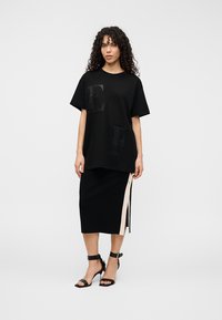 Μαύρο υπερμεγέθες t-shirt με ανάγλυφα γράμματα "E" και "F" στο εμπρόσθιο μέρος, συνδυασμένο με μαύρη εφαρμοστή φούστα με μπεζ πλαϊνό άνοιγμα και μεταλλικά στοιχεία.