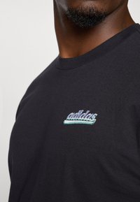 Čierne bavlnené tričko s krátkymi rukávmi a okrúhlym výstrihom. Obsahuje vyšívané logo "adidas" v tyrkysovej a striebornej farbe.