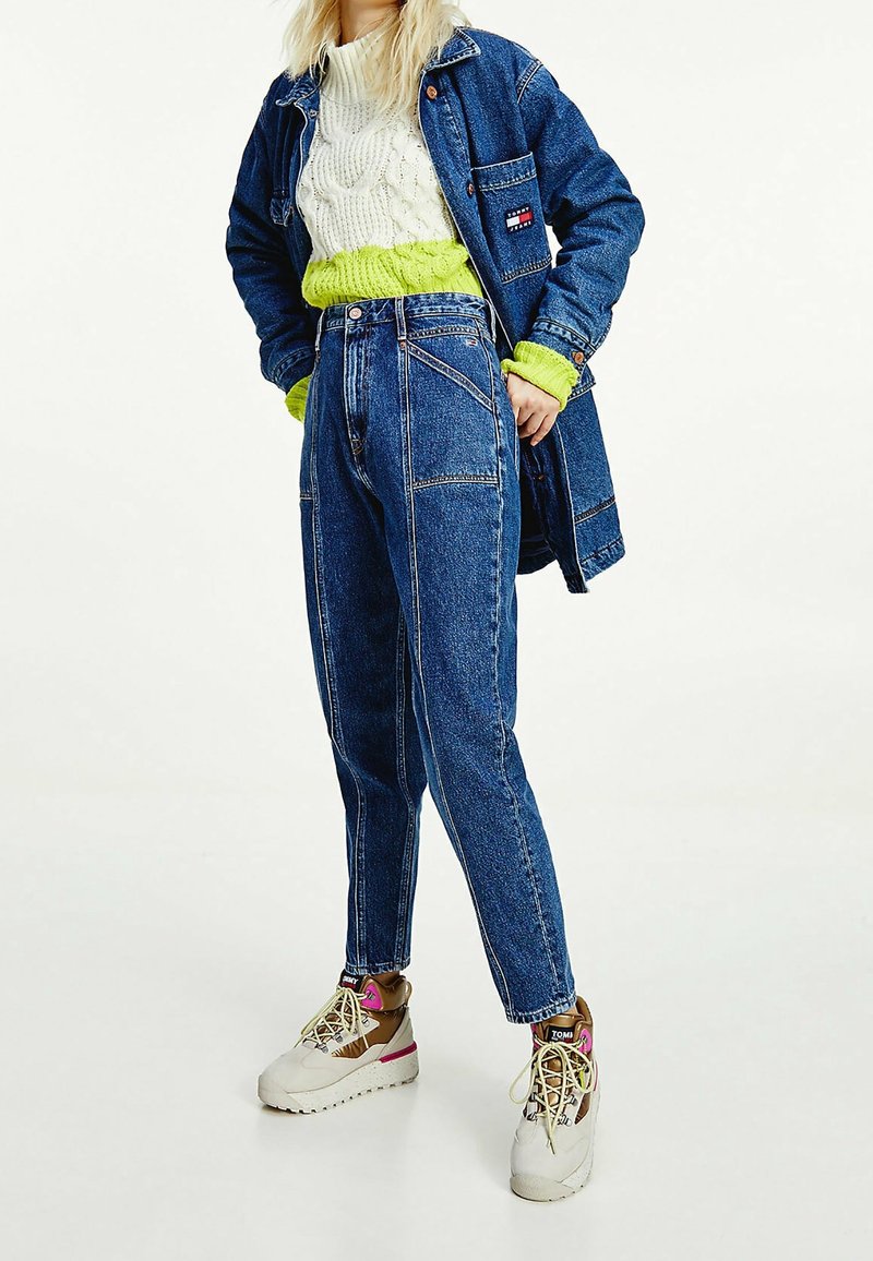Veste en denim, bleu clair ; pull en tricot blanc à cable superposé avec du vert vif ; jean bleu taille haute ; sneakers beiges avec des accents roses.