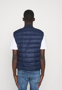 Hombre de espaldas vistiendo un chaleco acolchado azul marino, una camiseta blanca de manga corta y jeans azules sobre un fondo claro y liso.