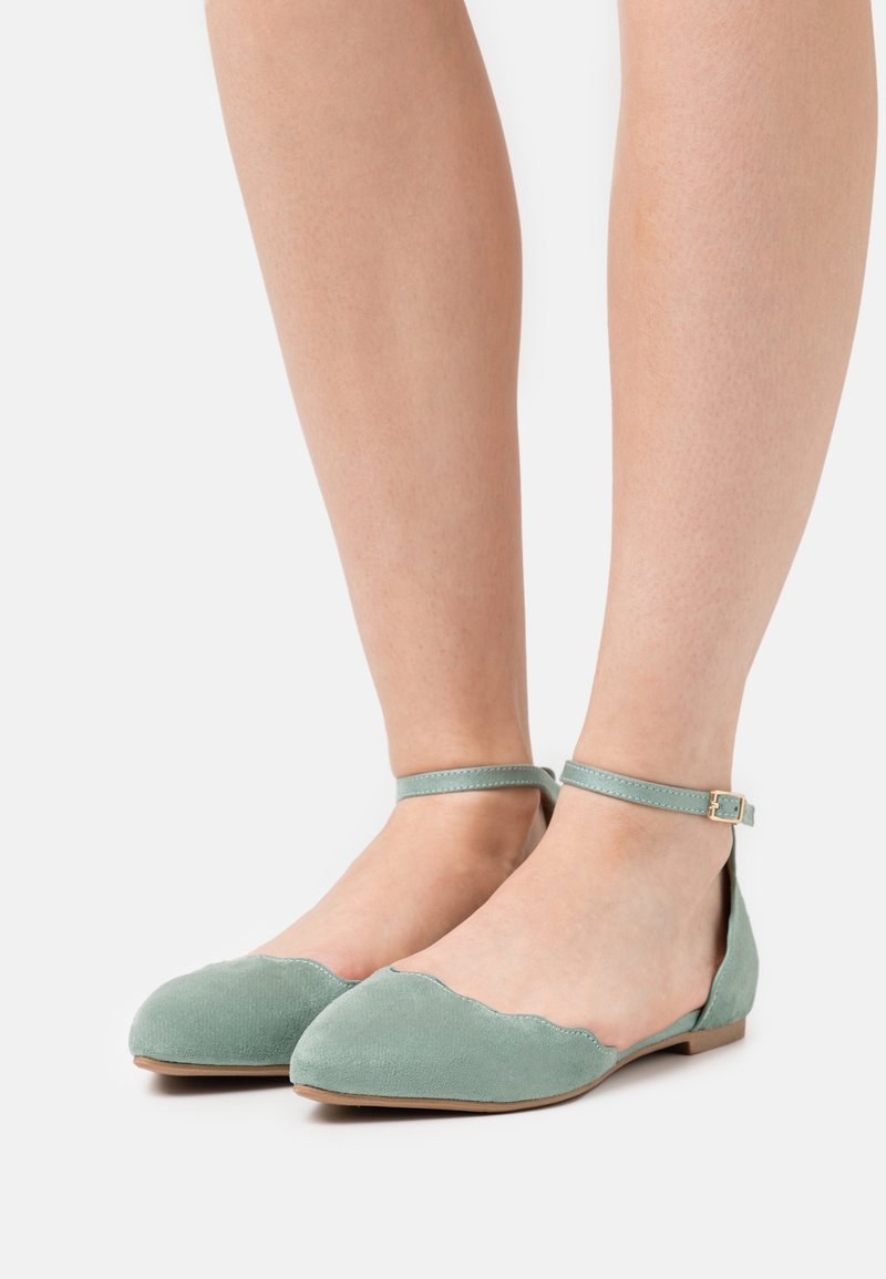 Zapatos planos de ante verde menta con bordes randados y una correa alrededor del tobillo, que presentan una pequeña hebilla dorada y un diseño de punta redonda.