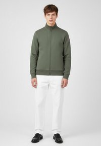 Olivgrön zip-up jacka med hög krage, texturerat tyg och ribbade mudd, i kombination med vita byxor och svarta loafers.