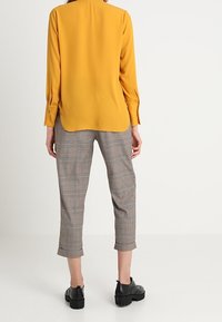 Senfgelbe Bluse mit langen Ärmeln, abgerundetem Saum und Knopfmanschetten, kombiniert mit grauen karierten, verkürzten Hosen und schwarzen Plateauschuhen.