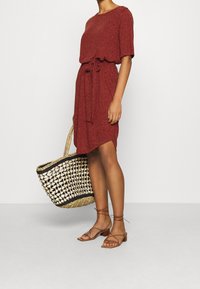 Rotes, kurzärmeliges Kleid mit einem Bindegürtel und einem hohen-senkrechten Saum. Braune, Riemchensandalen und eine geflochtene Tasche mit schwarzem Rand runden den Look ab.