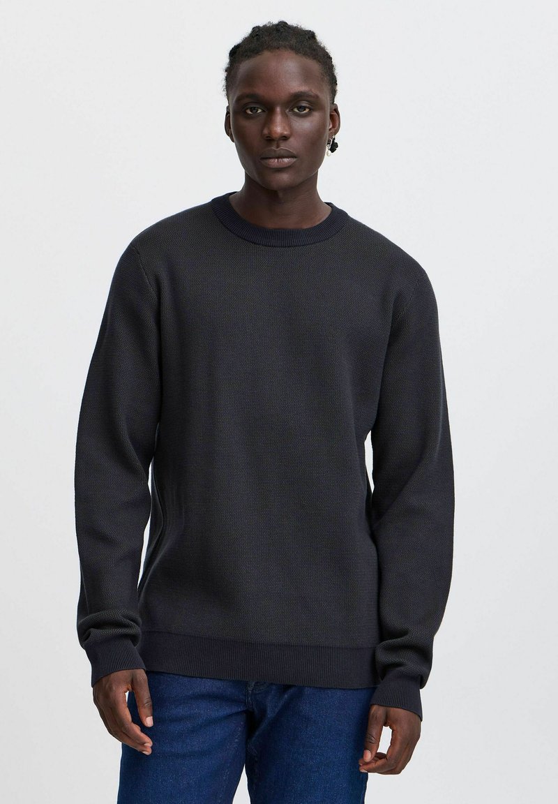 Dunkelblaues Strickpullover mit strukturiertem Design, Rundhalskragen und langen Ärmeln. Mit Rippbündchen und Saum. Kombiniert mit blauen Jeans.
