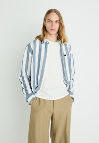 Blue and white striped button-up shirt with a relaxed fit, paired with a white T-shirt and beige wide-leg trousers. Smooth fabric texture.