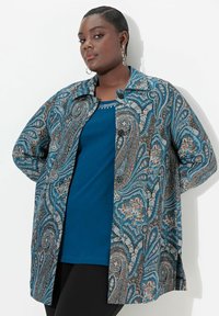 Modrá košile s paisley potiskem, s dlouhými rukávy, límečkem a kontrastním pevným modrým topem pod ní. Černé kalhoty.