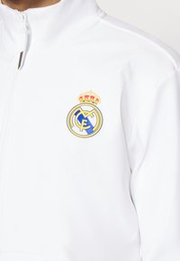 Veste blanche zippée avec l'écusson du club de football du Real Madrid, présentant une couronne et des lettres jaunes et bleues entrelacées sur la poitrine.