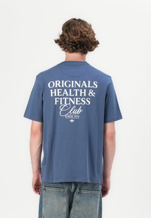 Blaues Baumwoll-T-Shirt mit kurzen Ärmeln, das auf der Rückseite weißen Text trägt: "ORIGINALS HEALTH & FITNESS Club seit 1972." Lässiger Schnitt.