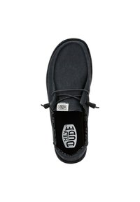 Zapato slip-on de lona negra con puntera redonda, detalle de cordones y un interior con patrón. Presenta una plantilla con marca y costuras en contraste.