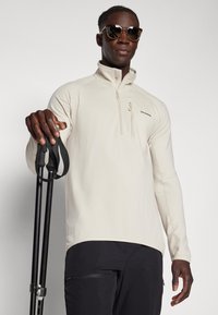 Pullover beige a mezza zip con texture a costine, maniche lunghe e logo Salomon. Il modello tiene in mano dei bastoni da trekking, indossa occhiali da sole e pantaloni neri.