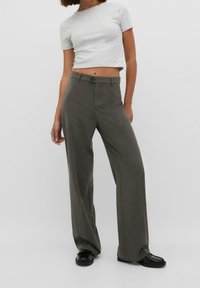 Grå cropped t-shirt, højtaljede, letvægtsstof. Mørke olivenfarvede brede bukser med forlommer og en subtil tekstur. Sorte sko.