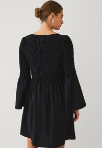 Zwarte jurk met smock-bodice, lange klokmouwen en een uitlopende rok. Gemaakt van een gestructureerde stof, met een schoon en simpel ontwerp.