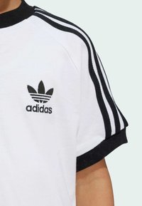 Biela tričko s čiernym golierom a manžetami, s čiernym logom Adidas a tromi čiernymi pásikmi na každom ramene. Mäkký bavlnený materiál.