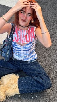 Bílé tílko s modro-červeným grafickým motivem, otevřený zadní díl. Tmavě modré džínsy s puntíky. Doplňky zahrnují černý obojek a kruhové náušnice.