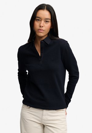 Vrouw met lang donker haar die een zwart poloshirt met lange mouwen en licht beige broek draagt, staat met handen achter haar rug.