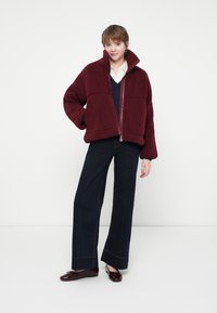 Persoană îmbrăcată cu o jachetă de fleece burgundy, cămașă albă, pulover bleumarin, blugi largi negri și pantofi burgundy stând în fața unui fundal alb.