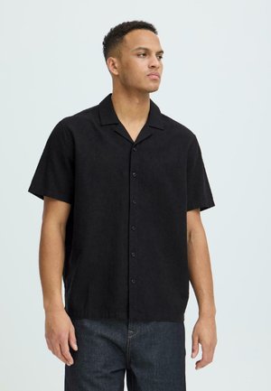 Jeune homme portant une chemise noire à manches courtes et un jean foncé, regardant sur le côté sur un fond clair uni.