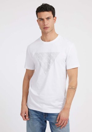 T-shirt en coton blanc avec un motif graphique triangulaire en gris. Manches courtes et col rond. Porté avec un jean en denim bleu.