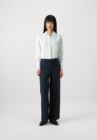 Weißes Hemd mit gerüschter Vorderseite und langen Ärmeln, kombiniert mit hoch taillierten navyblauen Hosen; figurbetonte Silhouette, glattes Material und spitze Schuhe.