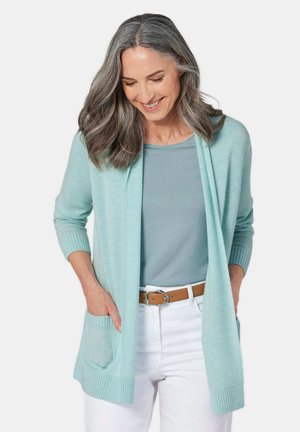 Cardigan bleu clair avec des poignets côtelés et une poche. Porté par-dessus un haut assorti, associé à un pantalon blanc et une ceinture marron. Texture lisse.