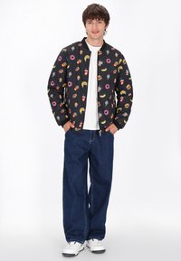 Giubbotto bomber nero con motivi colorati a tema cibo, abbinato a jeans blu larghi e sneakers bianche. Tessuto morbido, zip frontale, vestibilità casual.