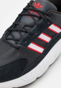 Sneaker med ovandel i svart nät med mörka mockadetaljer, tre vita och röda ränder samt en vit gummisula med texturerad detaljer.