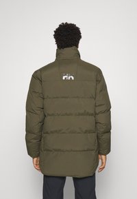 Olivově zelená puffer bunda s vysokým límcem, která má horizontální prošívání a odrazový detail loga na zadní straně. Hladká textura, délka po kolena.