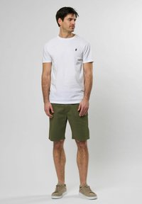 Camiseta de algodón blanca con cuello redondo y pequeño logotipo negro, combinada con pantalones cortos verdes y zapatillas beige, con un corte relajado.