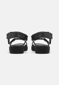Des sandales noires avec une tige en cuir lisse, des sangles réglables et une semelle épaisse et texturée, ornées de détails en métal doré.