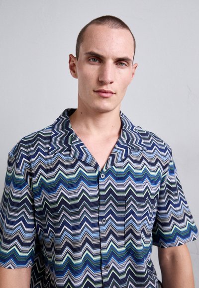 Ragazzo giovane con capelli corti che indossa una camicia a maniche corte con bottoni a zigzag blu, verde e nera su uno sfondo semplice.