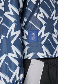 Modrá vzorovaná bunda s bílým zigzagovým designem, obsahující logo adidas, elastické manžety a detail s kapsou na zip.