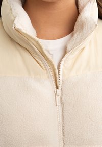 Veste en polaire avec un corps doux et texturé de couleur crème, une partie supérieure beige pâle et une fermeture éclair à l'avant. Caractérisée par un col haut et une fermeture éclair lisse.