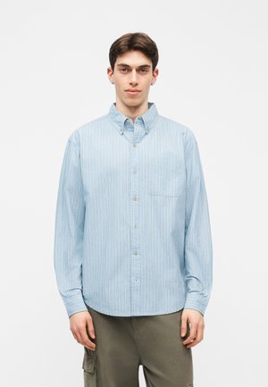 Νεαρός άνδρας με ανοιχτό μπλε ριγέ πουκάμισο και χακί παντελόνι cargo, που στέκεται μπροστά σε λευκό απλό φόντο.