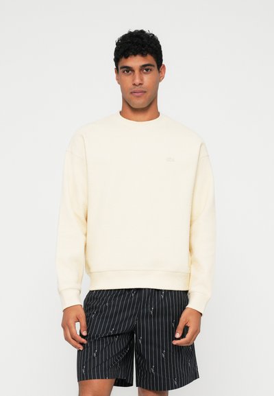 Sweatshirt crème avec des poignets et un ourlet côtelés, arborant un logo discret. Associé à un short noir à rayures, présentant un style décontracté.